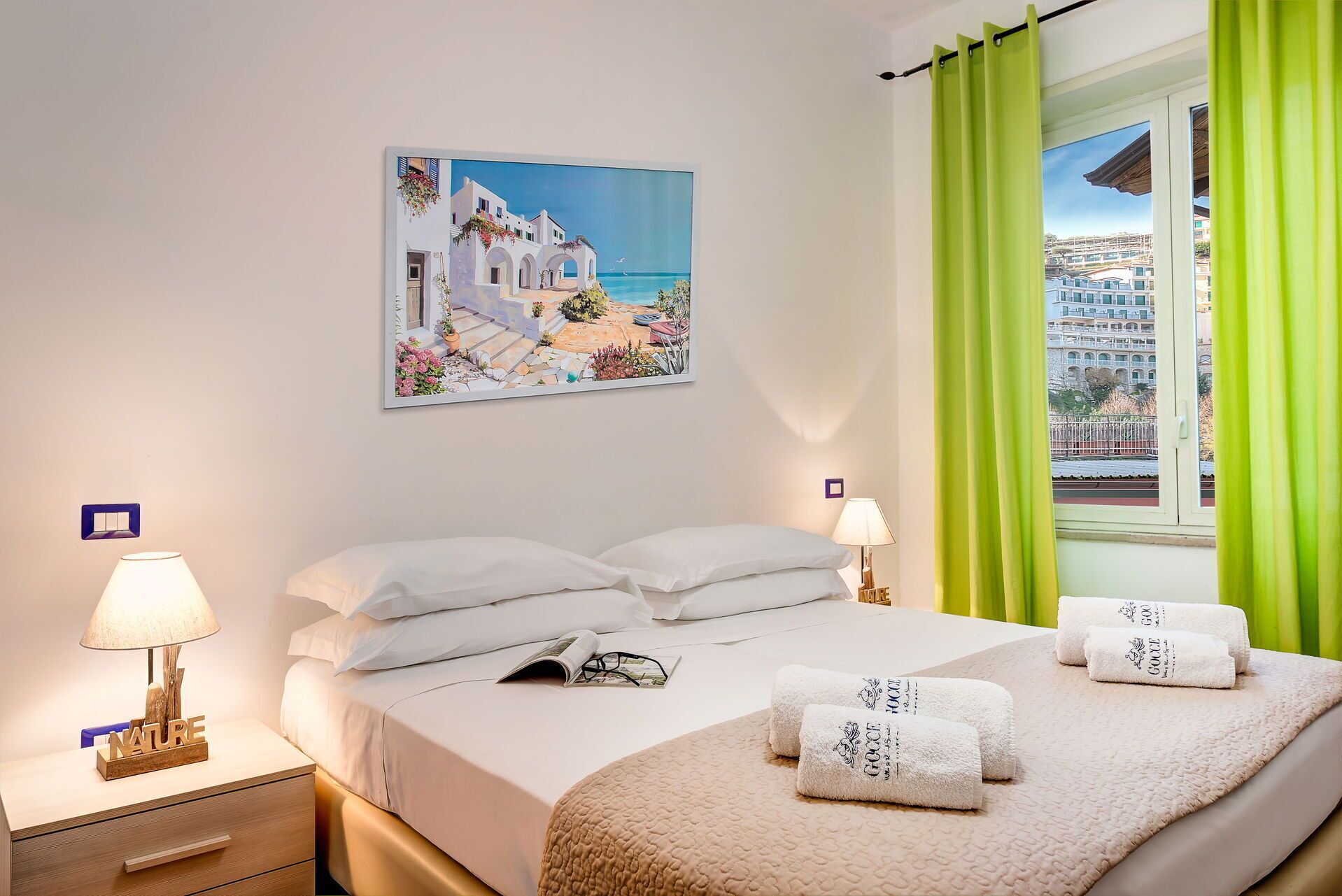 Ein Hotelzimmer mit einem Bett, zwei Nachttischlampen, einem Nachttisch, einem Fenster mit grünen Vorhängen und einem Gemälde einer Küstenlandschaft.