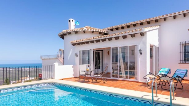 A poolside area with a clear view of the ocean, a covered patio with outdoor seating, and a white building with a tiled roof.