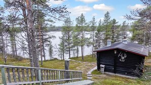 Una cabaña con terraza de madera y barandilla metálica, rodeada de árboles y un lago.