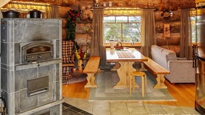 Interior rústico de una cabaña con chimenea de piedra, mobiliario de madera y un amplio ventanal con vistas al exterior.