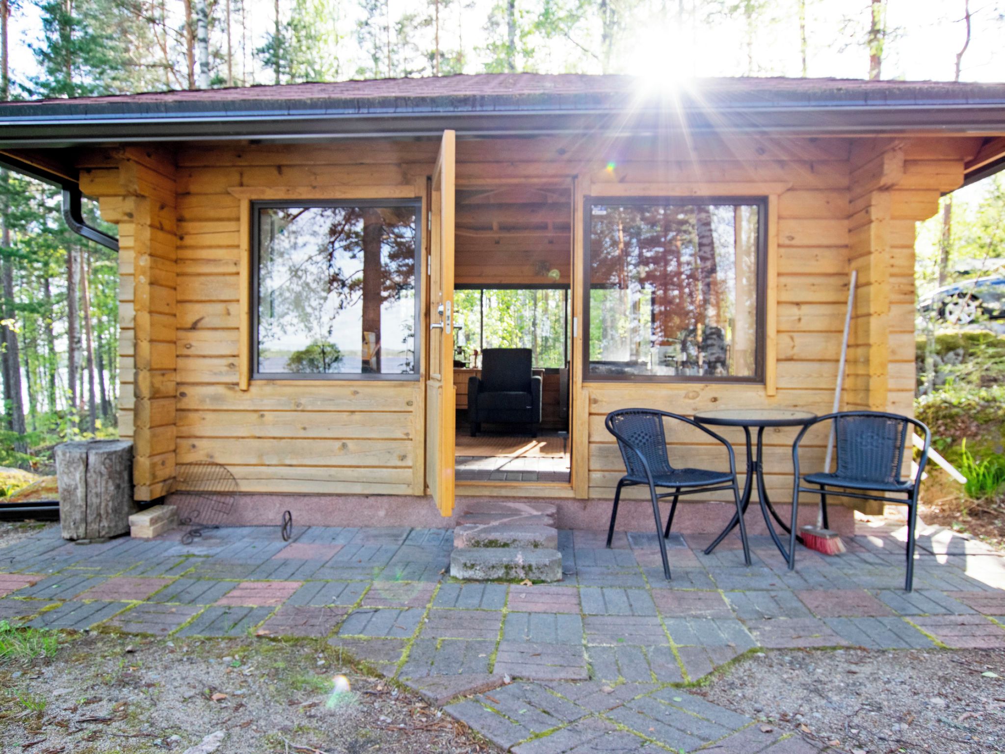 Eine Holzhütte mit großen Fenstern, einer Terrasse mit zwei Stühlen und Blick auf den Wald.