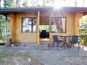 Eine Holzhütte mit großen Fenstern, einer Terrasse mit zwei Stühlen und Blick auf den Wald.