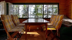 Interior de cabana com mesa e cadeiras de madeira, janelas amplas com vista para uma floresta e um tapete de palha no chão.