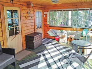 Het interieur van een blokhut met een geruit vloertje, een eettafel, stoelen, een open haard en een groot raam met uitzicht op het bos.
