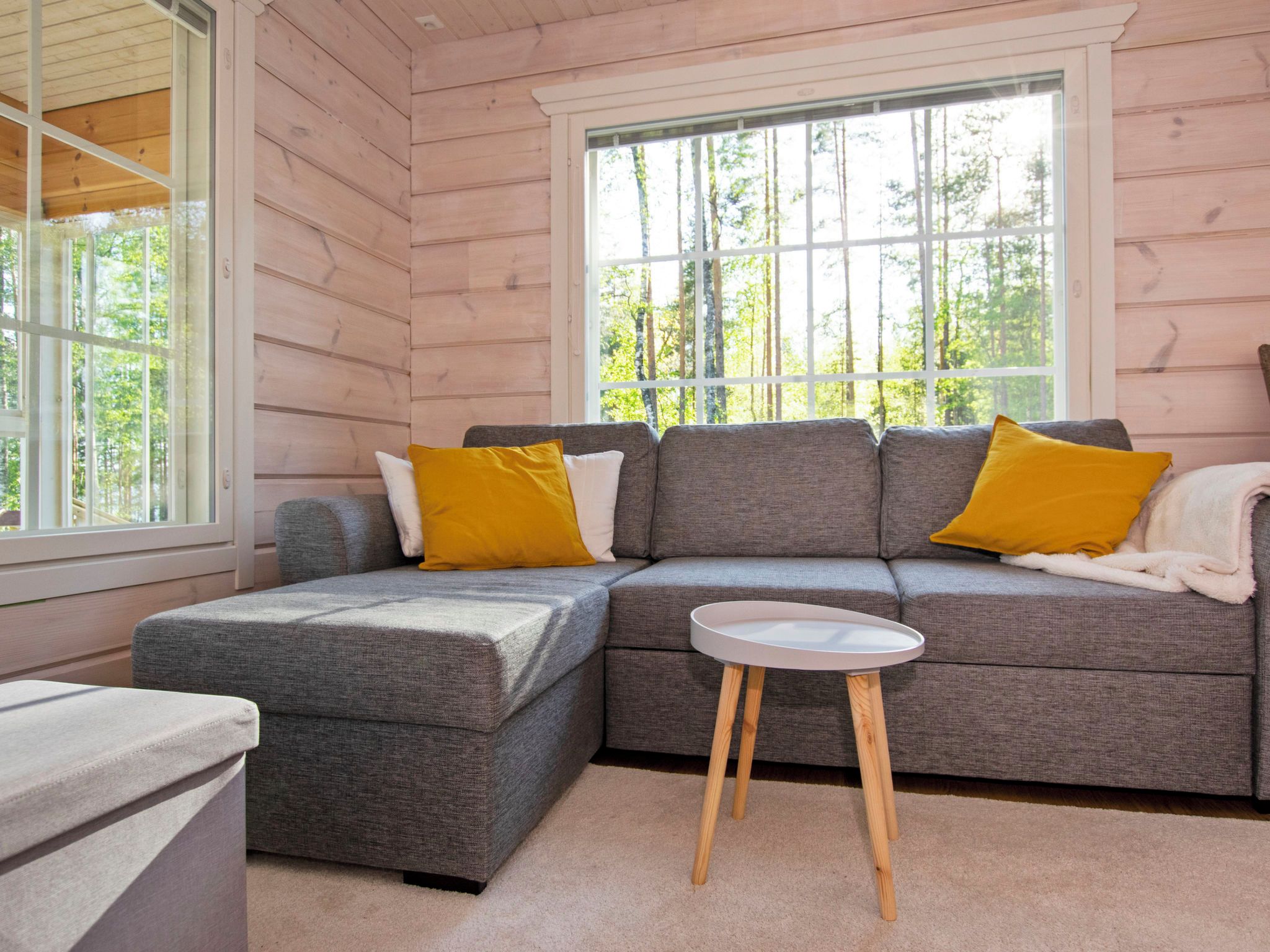 A cozy cabin interior with a gray sectional sofa, two yellow pillows, a small white table, and a window with a view of trees.
