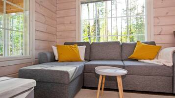 A cozy cabin interior with a gray sectional sofa, two yellow pillows, a small white table, and a window with a view of trees.