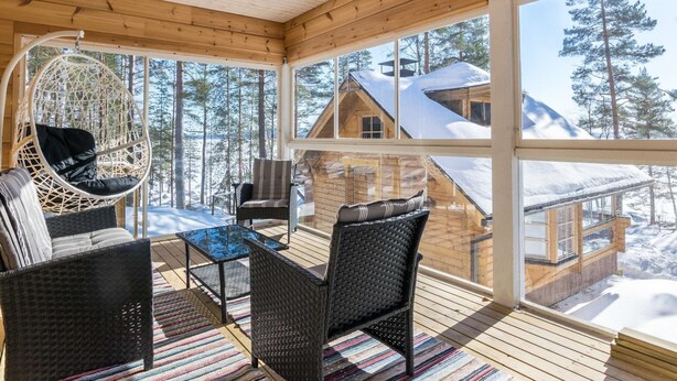Um deck de madeira com móveis de vime, uma cadeira suspensa e vista para uma cabana coberta de neve.
