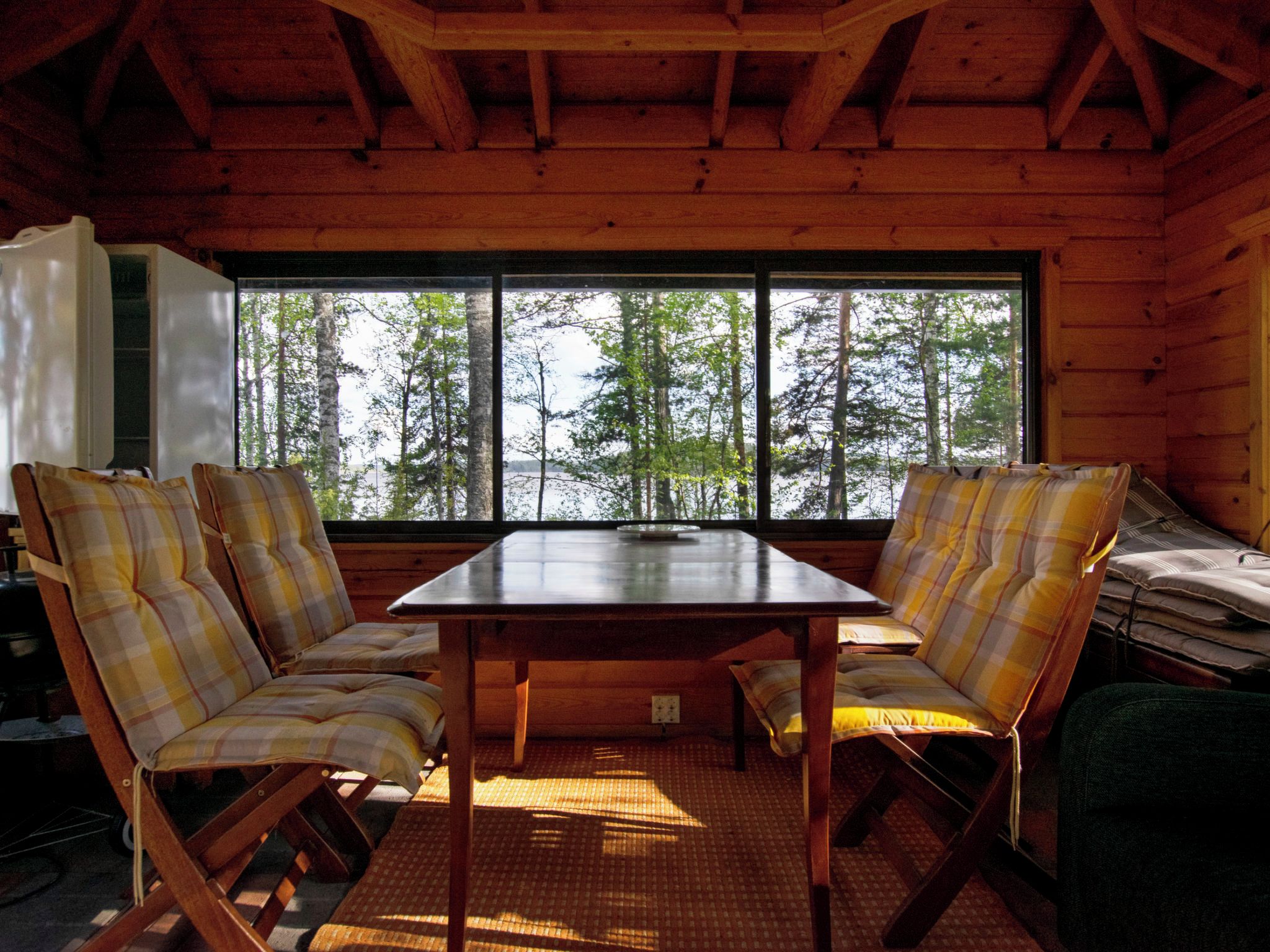 Ein Interieur einer Holzhütte mit einem großen Fenster, zwei karierten Sesseln, einem Tisch und Blick auf Bäume.