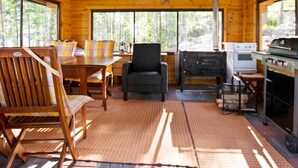 Interior de cabana com mesa de jantar de madeira, cadeiras, poltrona preta, cozinha com fogão e grelha, e amplas janelas que permitem entrada de luz natural.