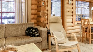 A log cabin interior with a sofa, a recliner, a dining table with chairs, and a window with curtains.
