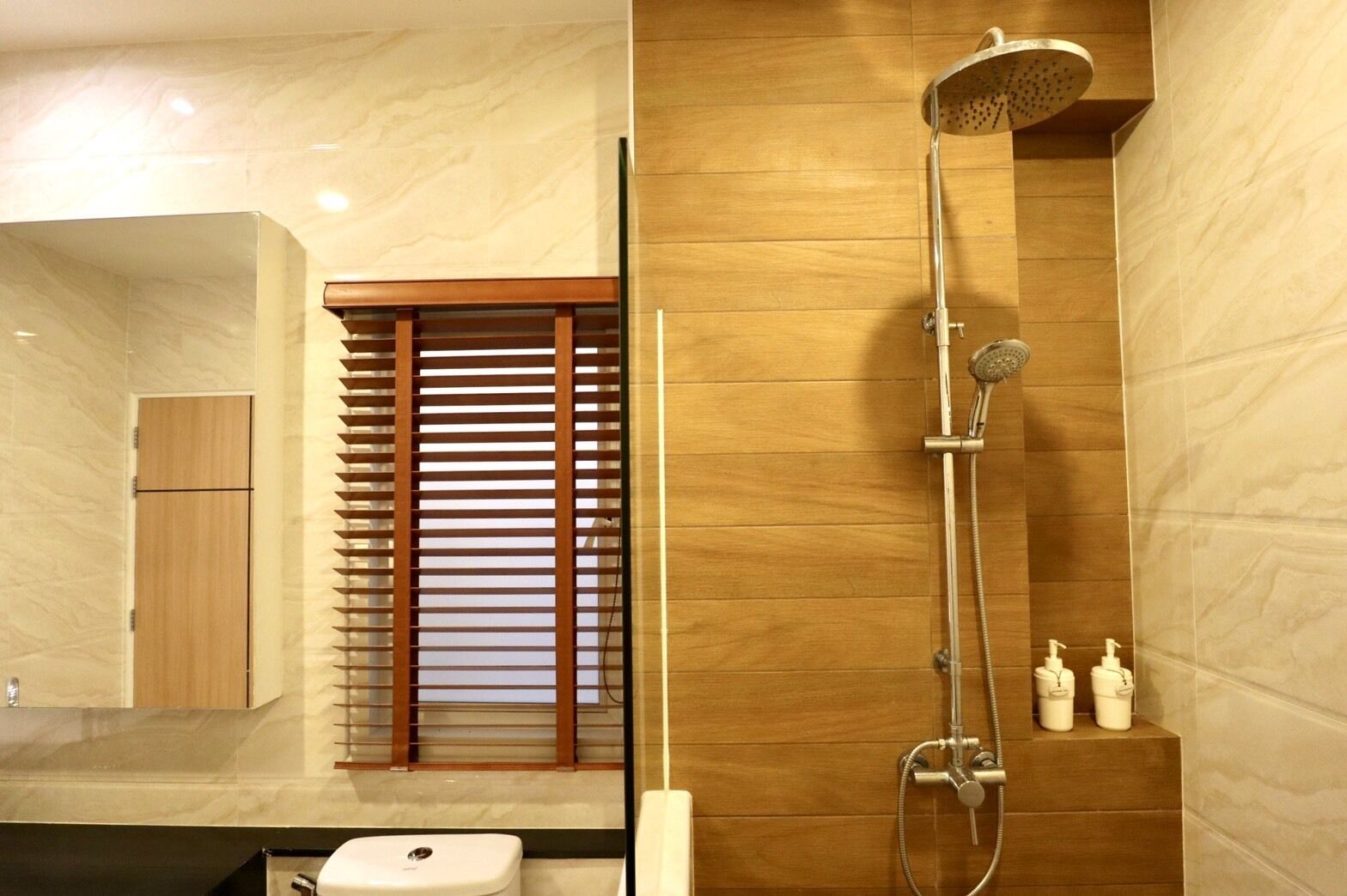 A modern bathroom with a wooden vanity, a toilet, a shower with a handheld showerhead, and a small shelf with toiletries.