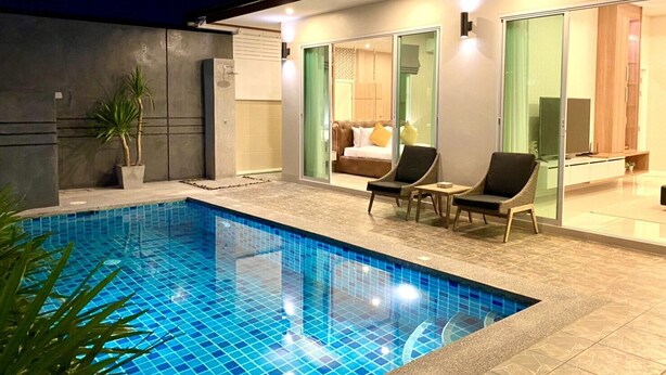 A modern indoor pool area with a clear pool, a seating area with two chairs, and a bedroom visible through a glass door. - Luxury Pool Villa A10  (Bang Lamung)