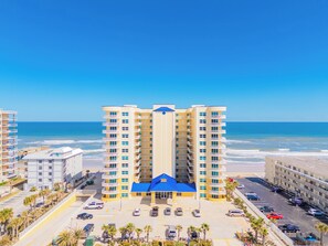 A multi-story hotel with a central blue-roofed building, surrounded by parking and palm trees, overlooking the beach. - Oceanfront-Heated Pool-Aruba #905 (Daytona Beach Shores)