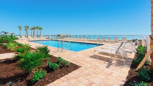 A pool area with lounge chairs and palm trees, overlooking a clear blue sky and ocean. - Oceanfront-Pool-Aruba #905-EV Charging (Daytona Beach Shores)