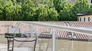 A balcony with a table and chairs, offering a view of a terracotta-tiled roof and lush greenery in the background.