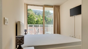 A hotel room with a large bed, a bedside table with a lamp, a TV mounted on the wall, and a window with a view of a cityscape and greenery.
