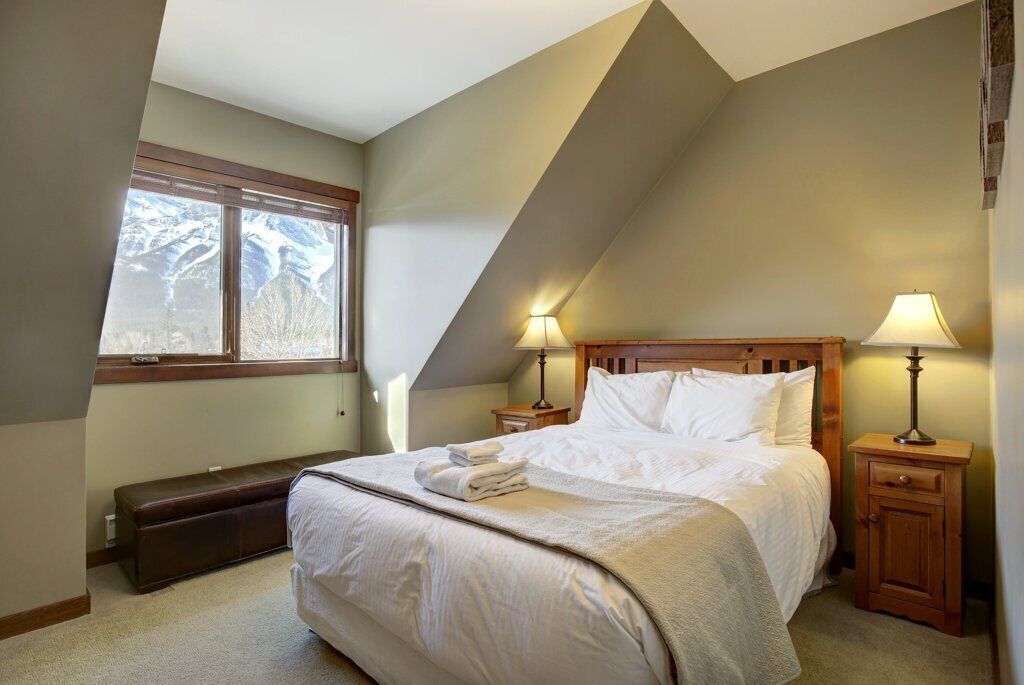 A bedroom with a large bed, two bedside lamps, a bench, and a window with a view of mountains.