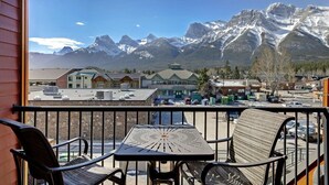 A balcony with a view of snow-capped mountains, a building with a green roof, and a parking area. - Canmore Place 312 | Downtown, Three Sisters Views (Canmore)