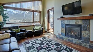 A living room with a fireplace, a television, and a large window with a mountain view. - Canmore Place 312 | Downtown, Three Sisters Views (Canmore)