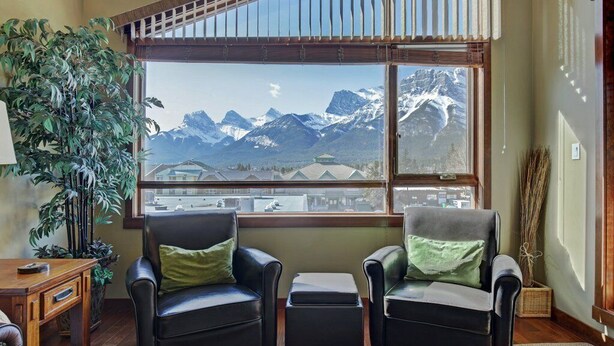 A room with two leather armchairs, a wooden side table, and a large window offering a mountain view. - Canmore Place 312 | Downtown, Three Sisters Views (Canmore)