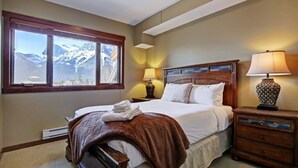 A bedroom with a large window offering a mountain view, a wooden headboard, and bedside lamps. - Canmore Place 312 | Downtown, Three Sisters Views (Canmore)