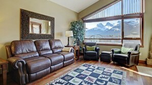 A living room with a leather sofa, two armchairs, a large window with a mountain view, and a patterned rug. - Canmore Place 312 | Downtown, Three Sisters Views (Canmore)