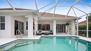 A covered outdoor swimming pool area with a glass ceiling, a seating area with a sofa and chairs, and a view of the surrounding landscape. - Picturesque Island Retreat ~Screened-In Lanai w/ Pool; Central Location w/Beach Gear & WiFi (Marco Island)