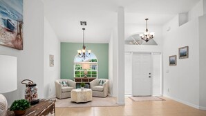 A spacious living room with two armchairs, a coffee table, and a chandelier. A large window with a view of greenery is visible. - Picturesque Island Retreat ~Screened-In Lanai w/ Pool; Central Location w/Beach Gear & WiFi (Marco Island)