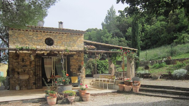 A rustic stone building with a stone roof, a wooden bench, and potted plants in front.