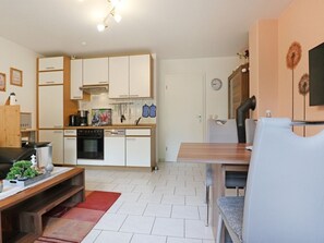 Uma cozinha compacta com armários brancos, uma área de refeições com mesa e cadeiras de madeira, e um pequeno espaço de estar com um banco.