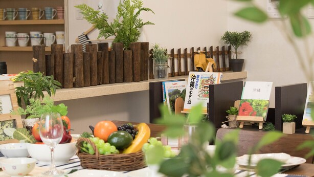 Ein Essbereich mit einer Holzwand, verschiedenen Früchten und Gemüsen, einem Weinglas sowie einer Auswahl an Büchern und kleinen Topfpflanzen.
