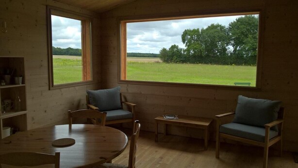 Habitación acogedora con detalles de madera, una mesa redonda, dos sillones y vistas a un campo verde a través de amplios ventanales.