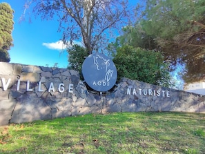 Un muro in pietra con la segnaletica 'VILLAGE AODE NATURISTE', immerso nel verde.