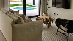 Un salon moderne avec un canapé, une table basse en bois, un téléviseur fixé au mur et une vue sur une piscine à l’extérieur.
