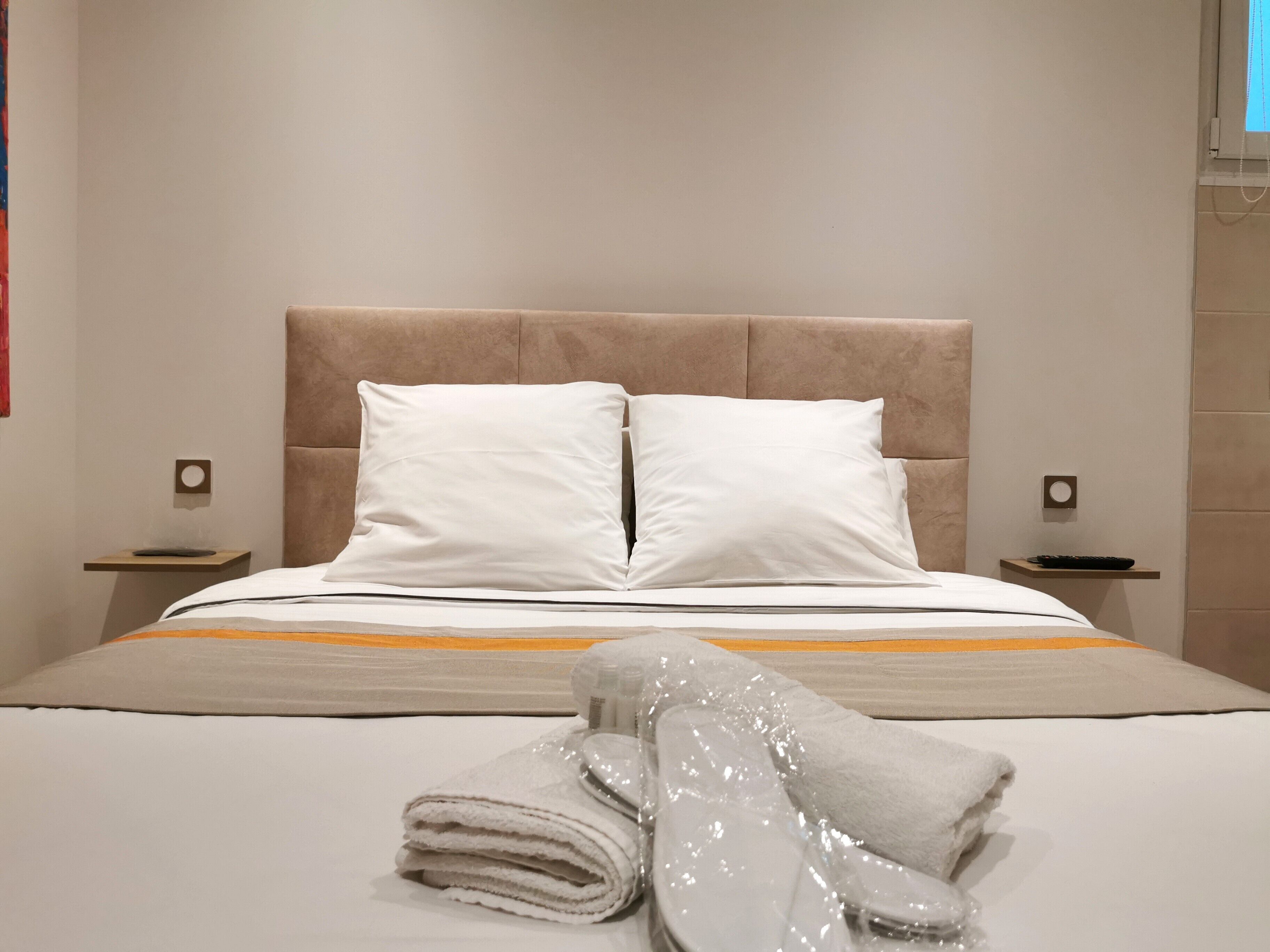 Un lit bien fait, avec des draps blancs et une tête de lit beige, agrémenté d’une serviette pliée et d’un objet emballé dans du plastique, posé sur le lit.
