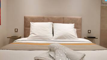 Un lit bien fait, avec des draps blancs et une tête de lit beige, agrémenté d’une serviette pliée et d’un objet emballé dans du plastique, posé sur le lit.
