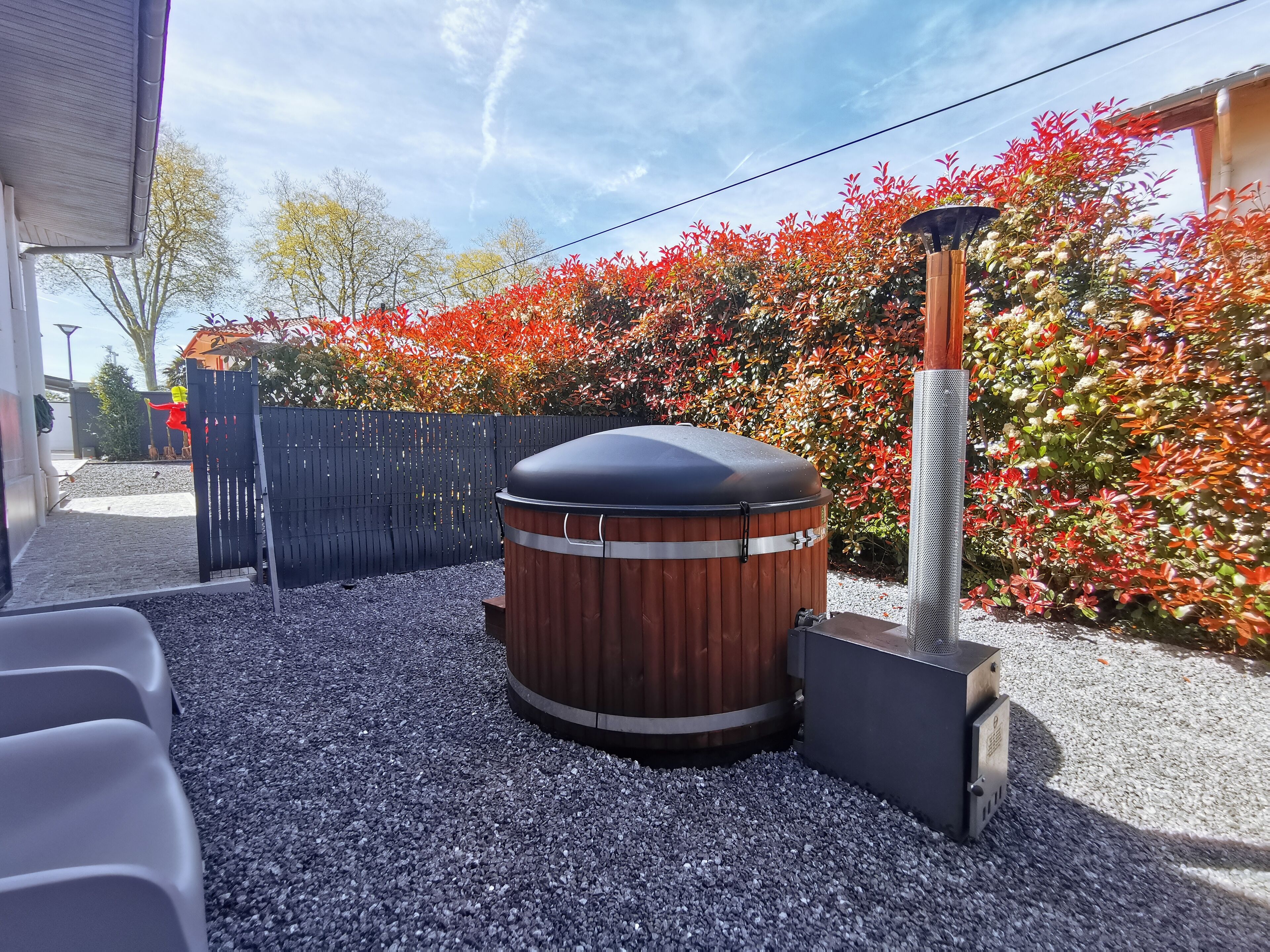 A hot tub with a wooden exterior and a metal base, surrounded by gravel and a wooden fence.