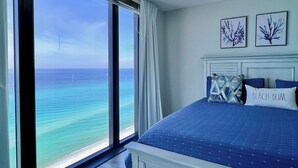A bedroom with a large window overlooking the ocean, a bed with a blue bedspread, and two framed pictures on the wall. - Picturesque Beachfront Condo- Spacious  Balcony, 2BD/2BA, Pool & Tiki Bar (panama city beach)