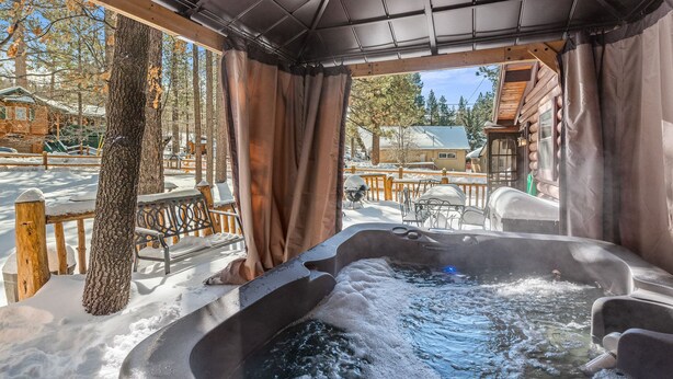 A hot tub area with a wooden deck, benches, and a view of a cabin and trees. - Adorable 1,500 sq ft log cabin, HOT TUB! Close to Lake, Village, & Forest! (Big Bear Lake)