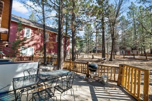 A wooden deck with metal chairs and a barbecue grill, surrounded by trees and a red building. - Log Cabin with Hot Tub | Across from Aspen Glen | Near Village & Lake (Big Bear Lake)