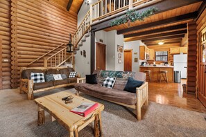 A log cabin interior with a living room, kitchen, and a staircase leading to a loft.