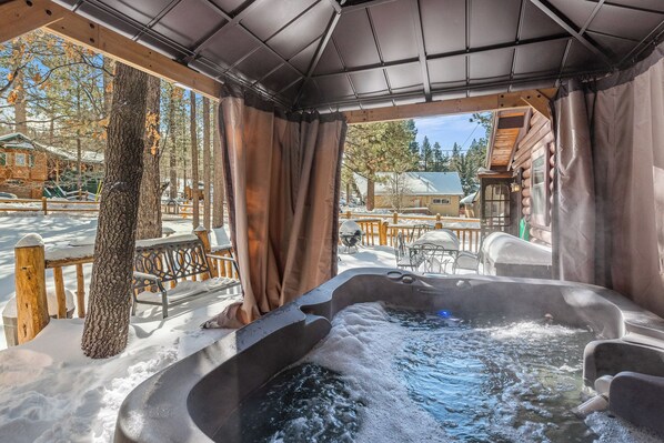 A hot tub area with a wooden deck, benches, and a view of a cabin and trees.