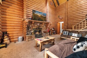 A log cabin interior with a stone fireplace, a wooden sofa, a coffee table, and a television mounted above the fireplace.
