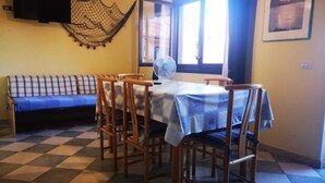A dining area with a checkered floor, a table with a blue tablecloth, wooden chairs, a sofa, a wall-mounted TV, and a window with curtains. - Apartment in La Maddalena (La Maddalena)