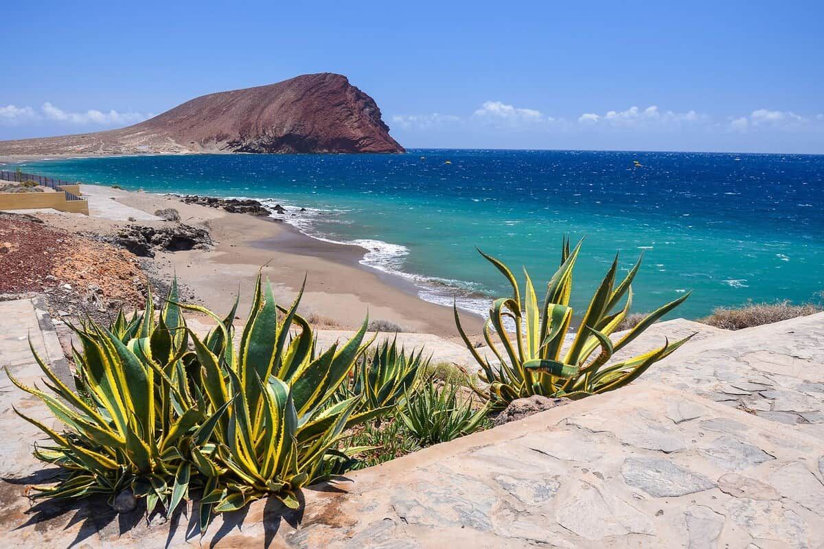 Eine Küstenlandschaft mit einem felsigen Strand, blauem Meer und einem markanten Berg im Hintergrund.