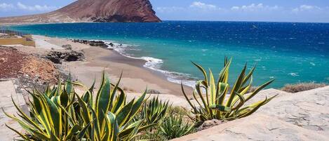 Eine Küstenlandschaft mit einem felsigen Strand, blauem Meer und einem markanten Berg im Hintergrund.
