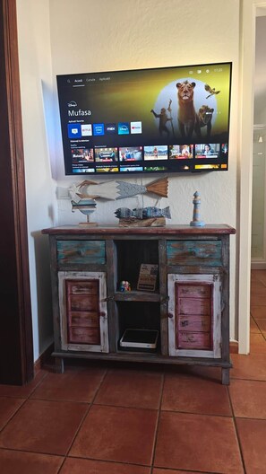 Un televisor montado en la pared muestra la interfaz de un servicio de streaming con diversas opciones de contenido. Debajo del televisor, hay un mueble rústico de madera con objetos decorativos y un pequeño dispositivo electrónico en su interior.
