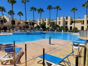 Una piscina con sillas de descanso, palmeras y un edificio con balcones al fondo.