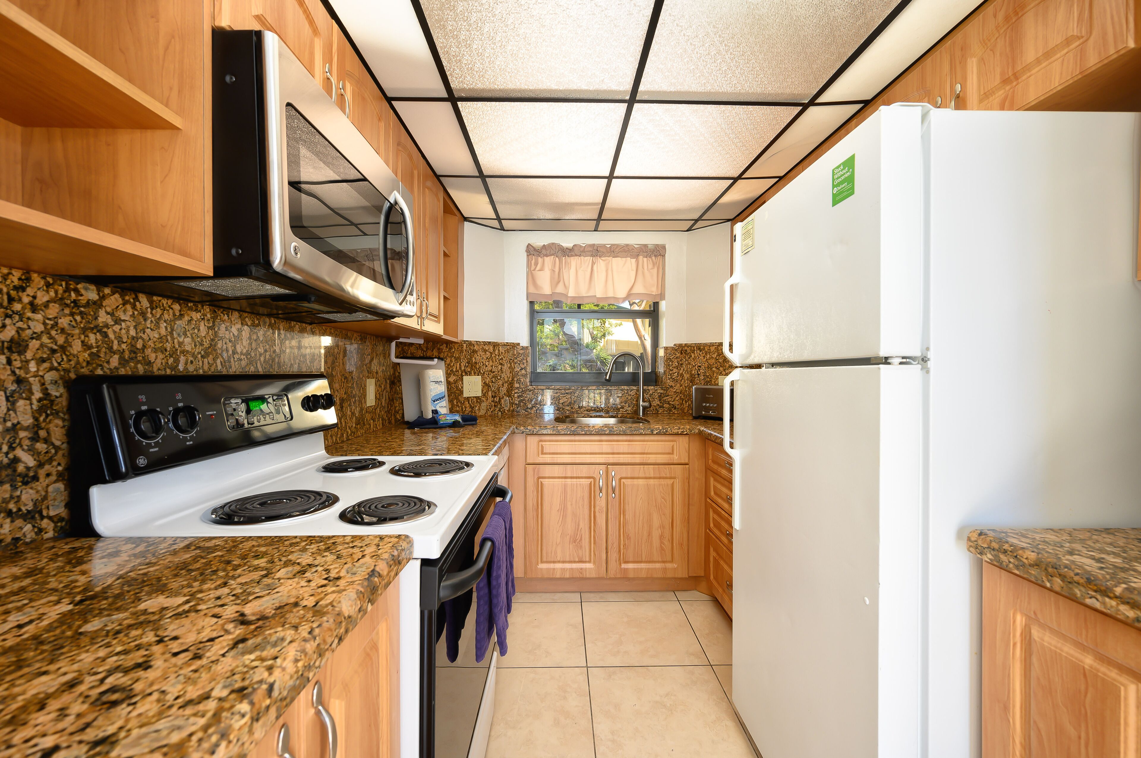 A kitchen with granite countertops, a white stove, a microwave, a refrigerator, wooden cabinets, and a window with a curtain.
