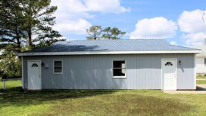 A single-story, light gray building with a sloped roof, two doors, and a small window. - Shore Bird - 1 BR Studio Cottage with open floor plan. NO LINENS PROVIDED (Chincoteague Island)